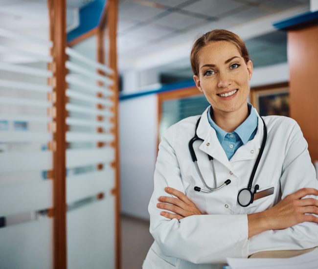 Smiling doctor standing with her arms crossed at the hospital and looking at camera. Copy space.